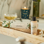 Bramley bubble bath Christmas cracker and bubble bath bottle on a wooden surface with a candle and glass in the background.