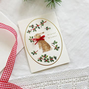 Christmas card with a rabbit and holly design on a white surface with a red ribbon.