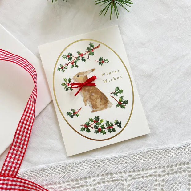 Christmas card with a rabbit and holly design on a white surface with a red ribbon.