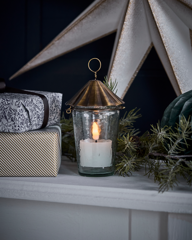 Decorative lantern with a candle on a shelf with a star-shaped object and greenery in the background.