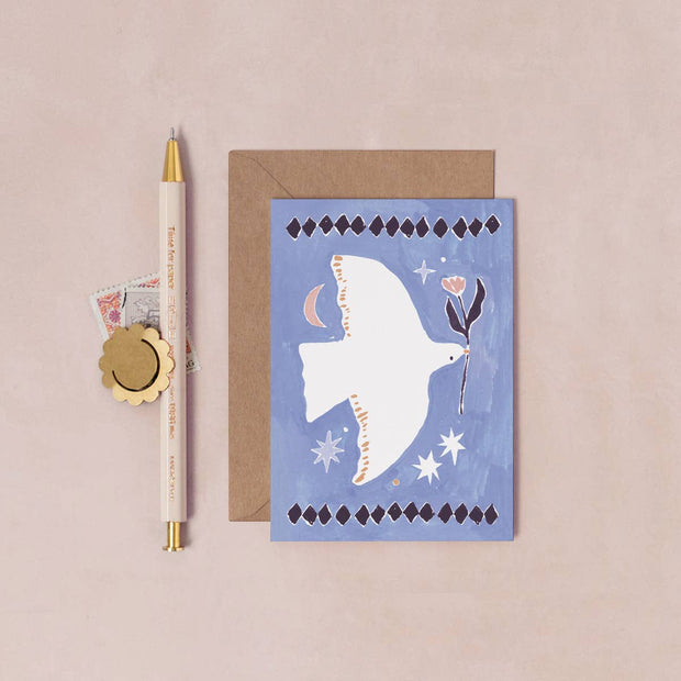 Greeting card with a dove illustration on a blue background, next to a pen and envelope on a beige surface.