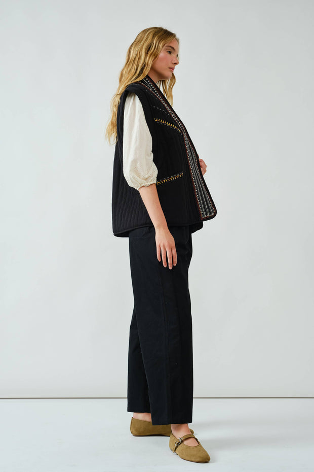 Woman wearing a black embroidered vest and dark pants on a white background