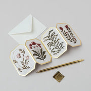 Hexagonal fold out cards with floral designs, a white envelope, and a gold pen on a light gray background