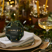 Rose Petal Bath Bombs Christmas Bauble-Bramley product packaging on a table with a glass of wine and decorative lights in the background