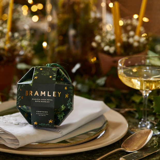 Rose Petal Bath Bombs Christmas Bauble-Bramley product packaging on a table with a glass of wine and decorative lights in the background