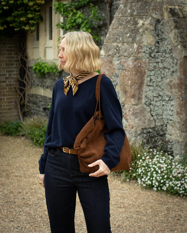 Woman holding a brown leather bag in front of a stone building with greenery.