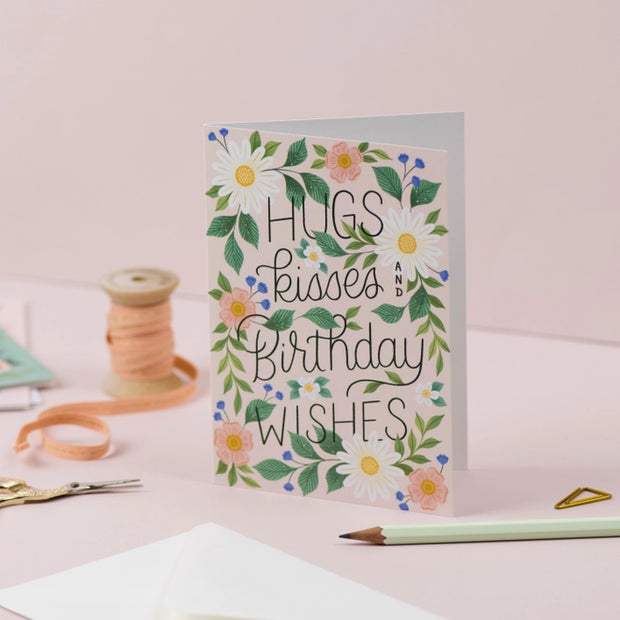 Fabulously floral, the caption on this female birthday card reads “Hugs, Kisses, Birthday Wishes” with handwritten lettering, surrounded by lush foliage, blooming flowers and forest green leaves all on a neutral background.