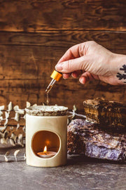 Hand using a dropper to add liquid into a small ceramic container with a lit candle, against a wooden background.