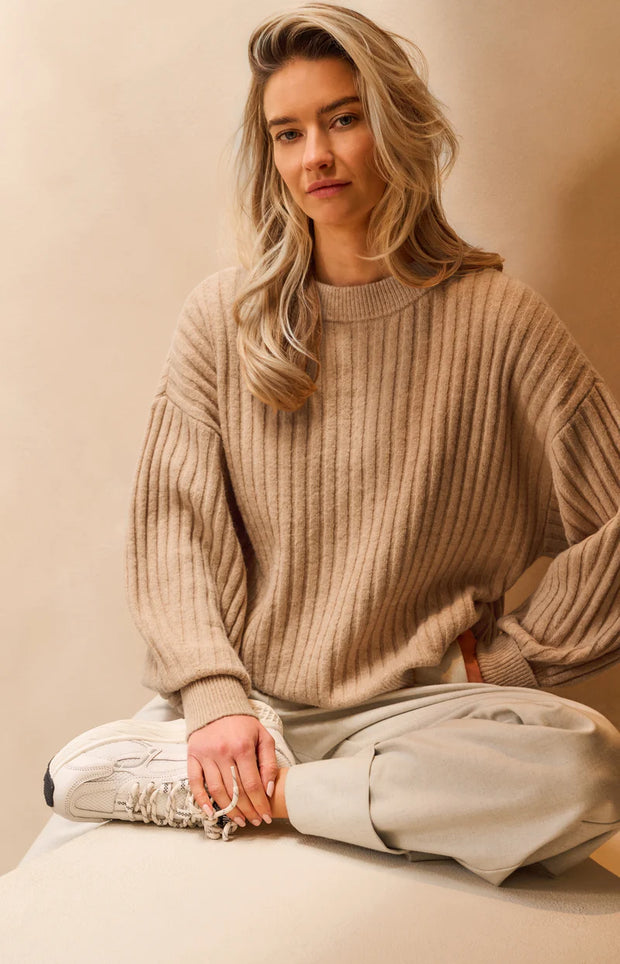 Woman wearing a beige sweater and light pants sitting on a white surface with a neutral background
