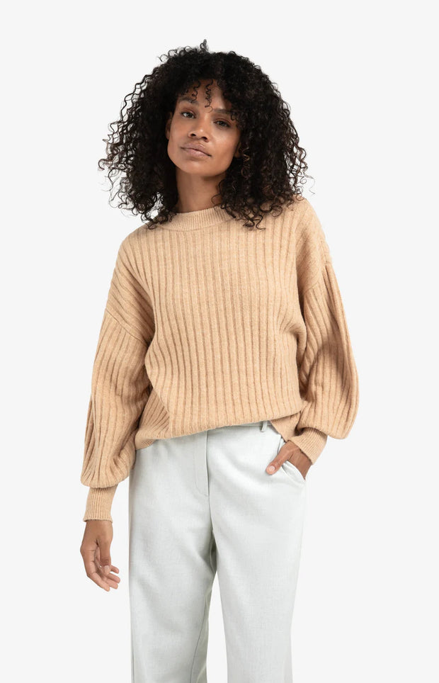 Woman wearing a beige ribbed sweater and white pants on a white background