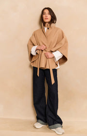 Woman wearing a beige cape over a white shirt with dark pants on a beige background