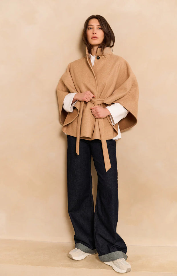 Woman wearing a beige cape over a white shirt with dark pants on a beige background