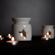 A white ceramic wax melt warmer with a star cut decal, accompanied by a tealight candle that is lit, placed on a dark surface with a blurred background.